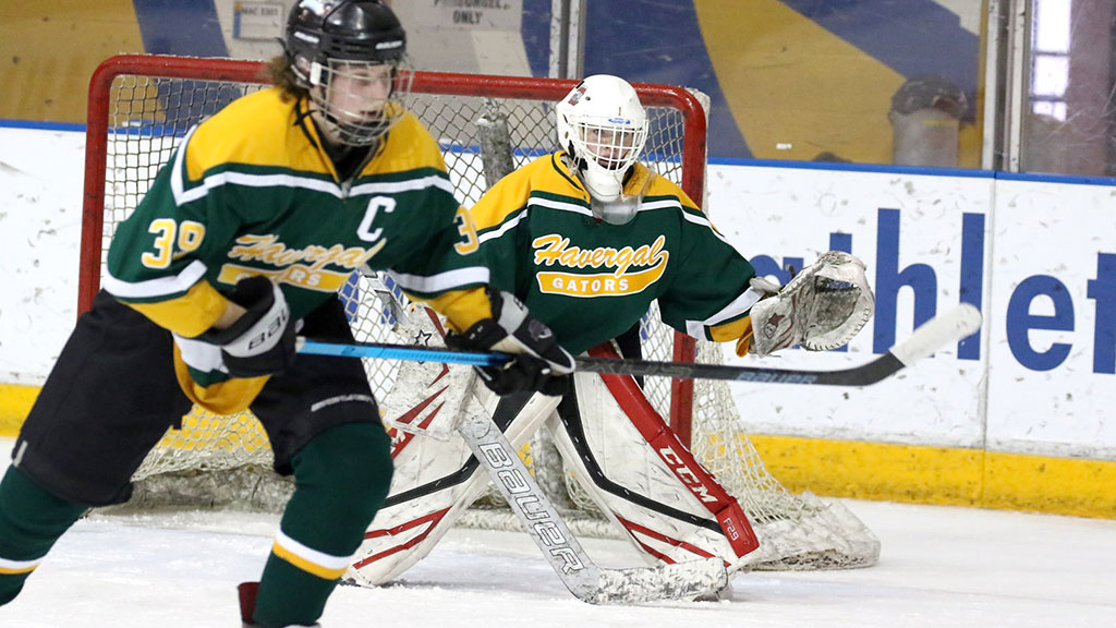 A goalie in a hockey net with a player beside her, both wearing green and gold.