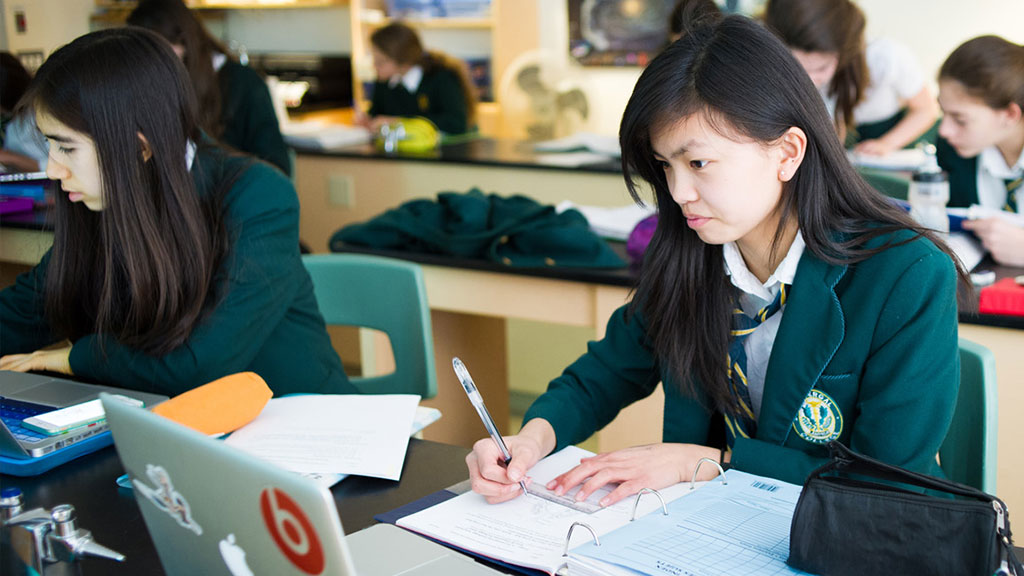 A student in a science class looking at a laptop while writing in a notebook.