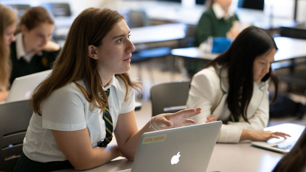 A student sitting in front of a laptop asking a question during class.