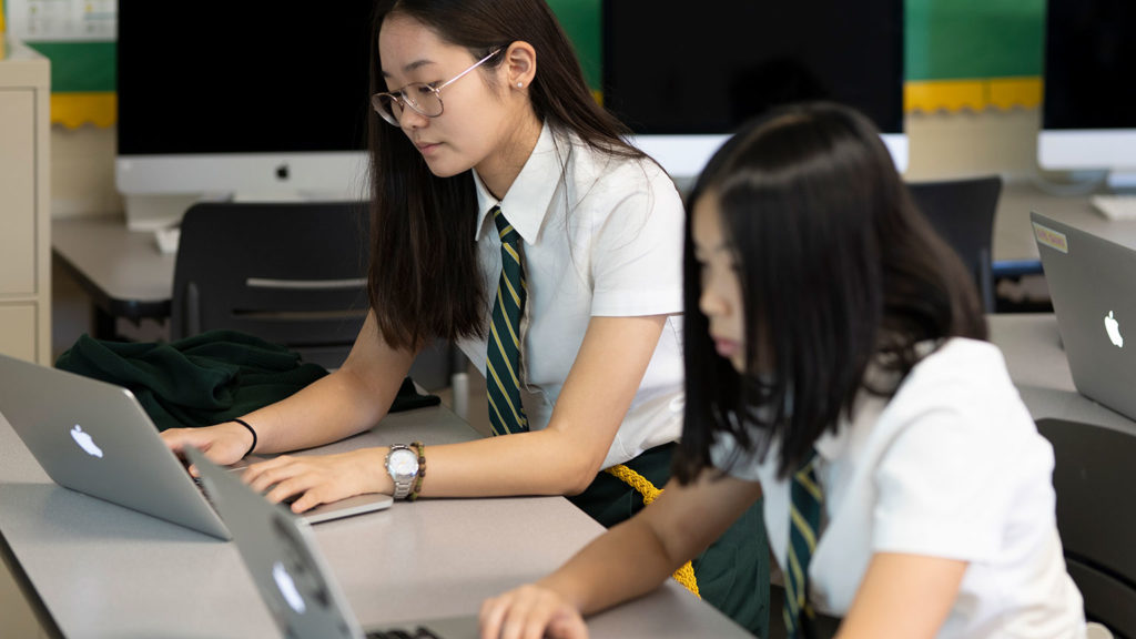 Two Senior School students type on laptops in a classroom.