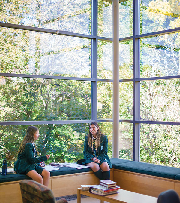 Two students sitting in front of windows with trees in the background.