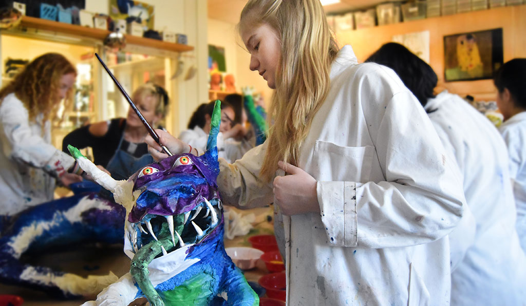 A student in an art smock paints a paper mache monster that she created.