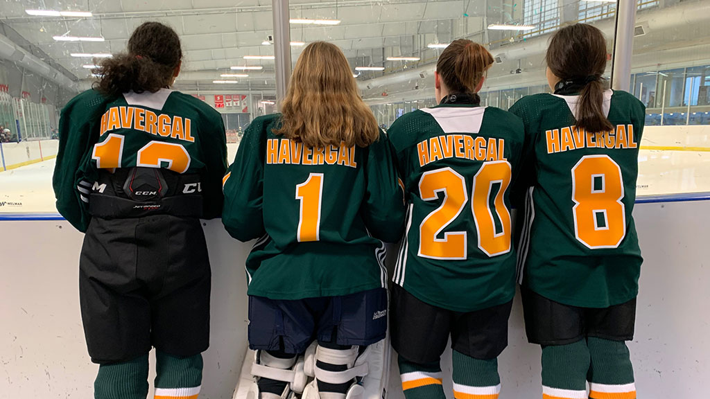 Four hockey players in Havergal uinforms stand together looking over the ice rink.