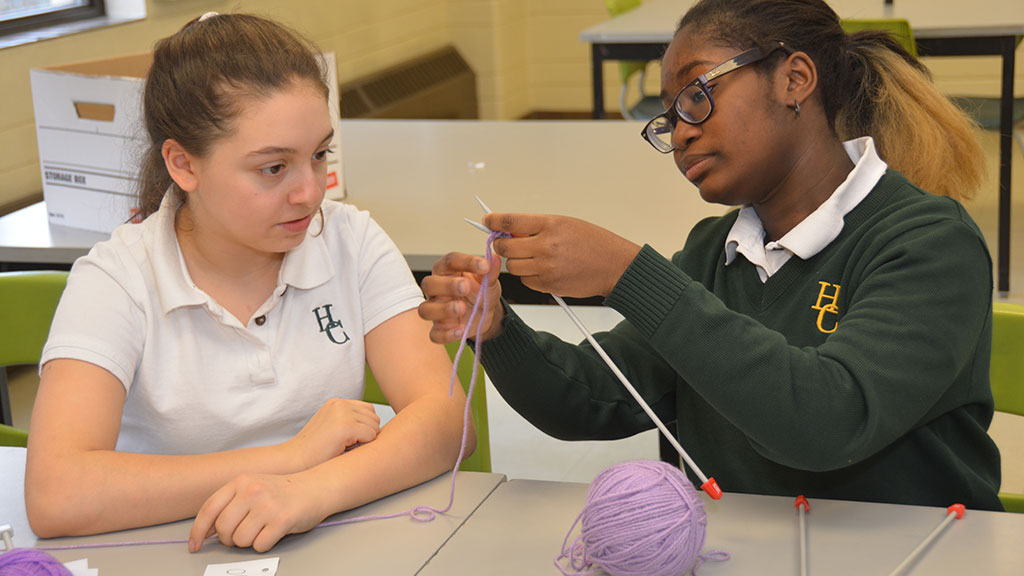 One student showing another how to knit, with purple yarn.