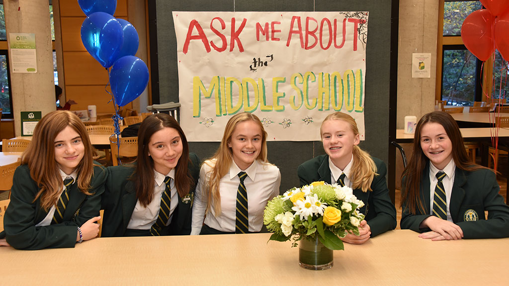 Five Middle School students sit at a table in front of a sign that reads Ask Me About the Middle School.