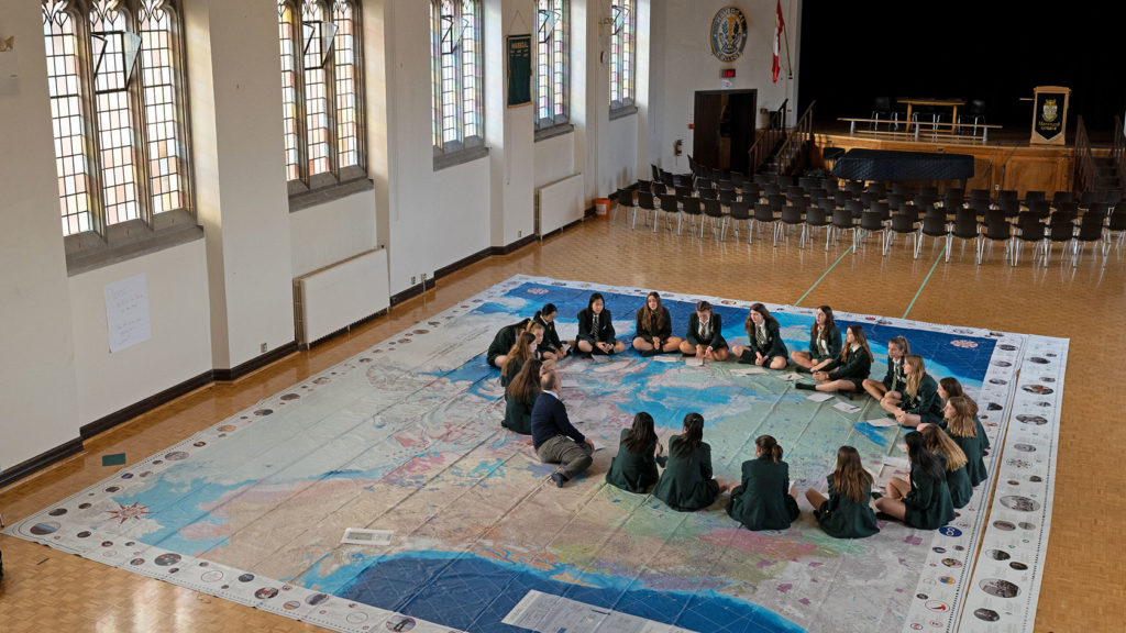 Students sit crossed-legged in a circle on a large map of Canada that is displayed on the floor of a hall.
