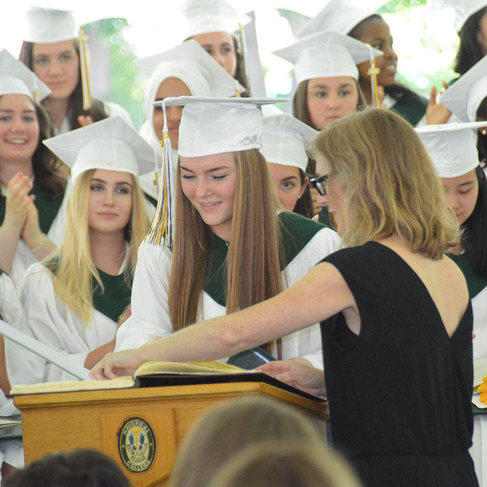 A student signs a book at a podium during graduation. Her peers stand behind her. All are wearing white caps and gowns with green sashes.