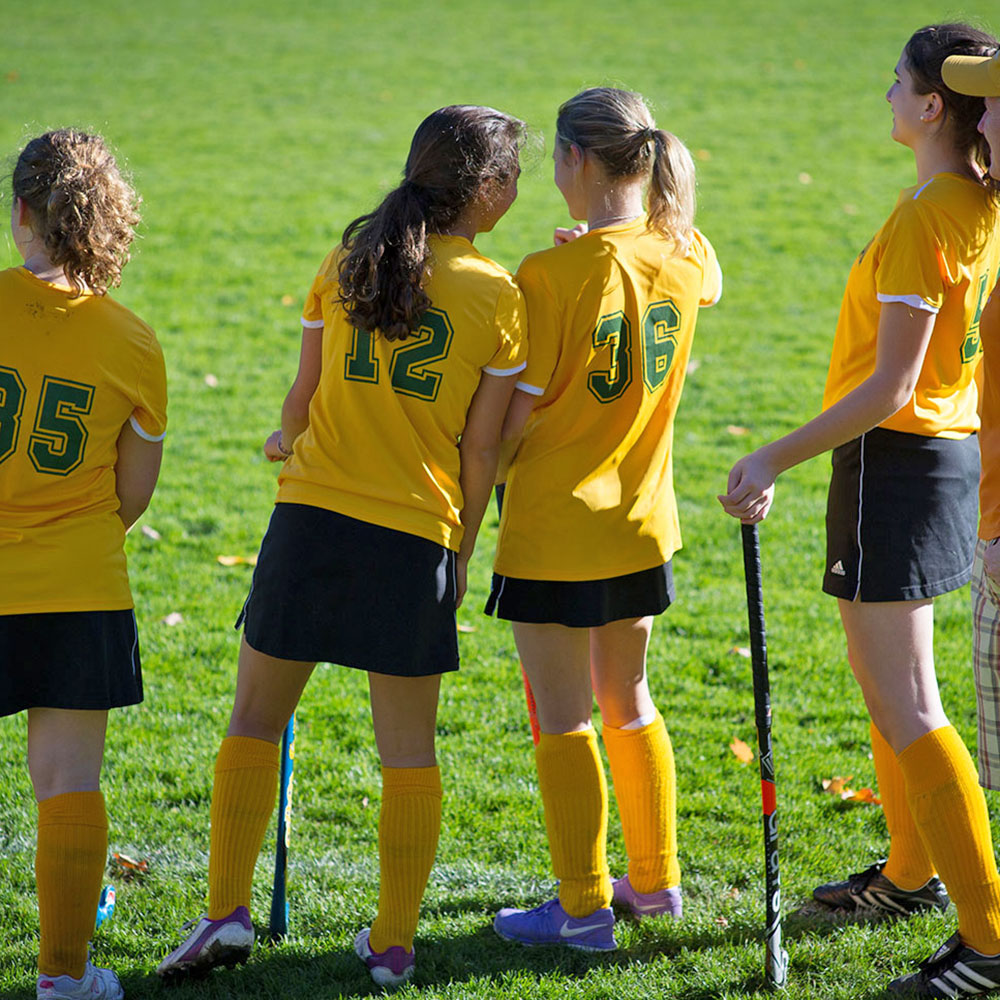 Four students in field hockey uniforms facing a field while in discussion with each other.