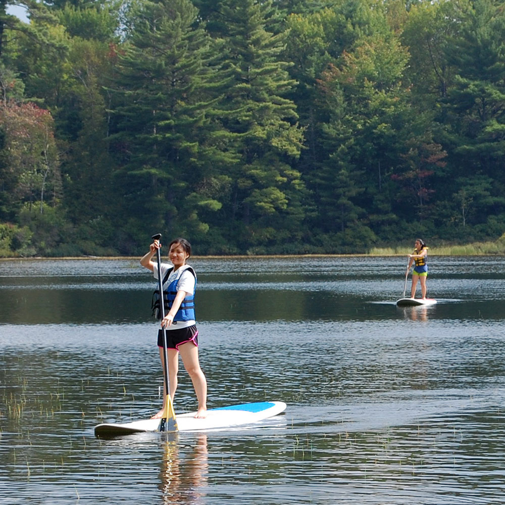 Two students stand up paddling on a lake in a forest.