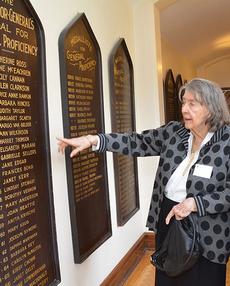 An elderly woman points to a list of names mounted on the wall of a school hallway.
