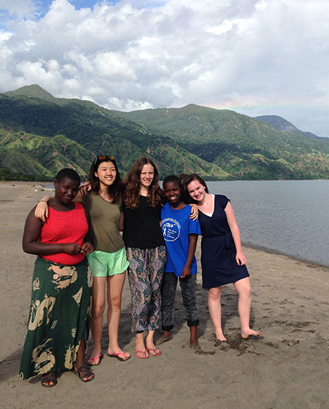 Students and their friends pose together on a beach in Tanzania with mountains and a rainbow in the background.