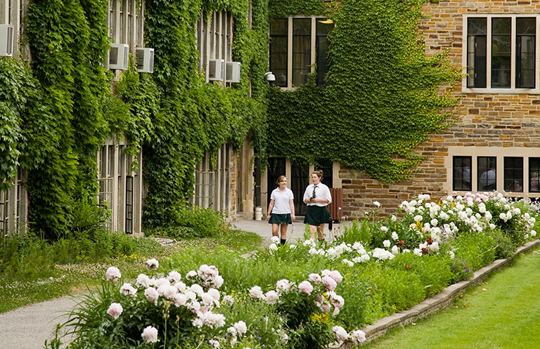 Two students walking down a path with an ivy-covered wall on the left and a garden on the right.