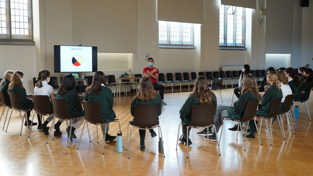 Students sit on chairs in a semi-circle while listening to Indigenous educator Mike Carlson.