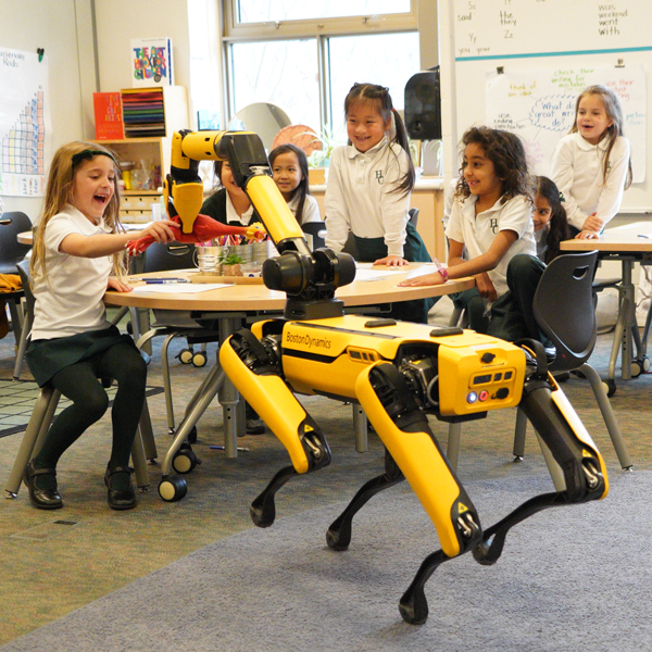 A yellow Boston Dynamics SPOT robotic dog hands a rubber chicken to a student in Grade 1.