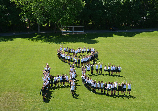 Students stand on Ratcliffe Field, where they combine to make a heart shape and the letters HC.