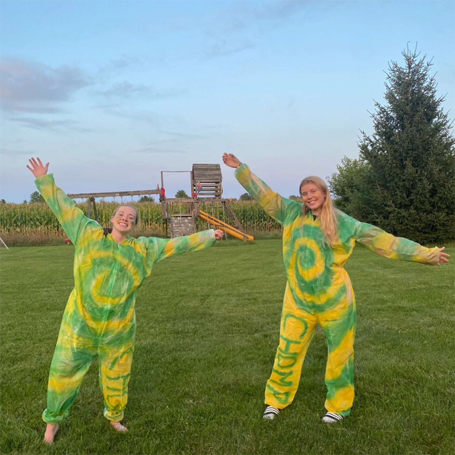 Kaitlyn and Paige standing in a sports field with their arms open wide as they wear yellow and green hazmat suits.